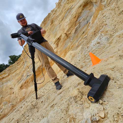 Person using a high-resolution metal detector on a sandy terrain with an orange cone nearby, that marks the scan line